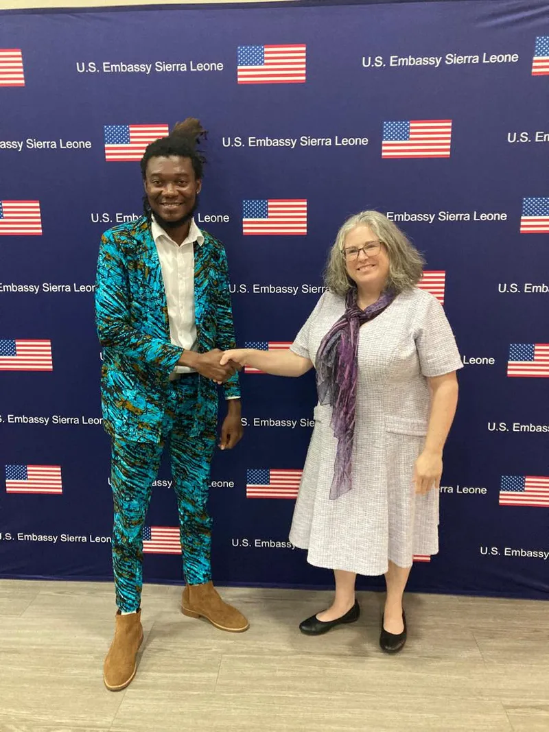 Joseph Jawah Kebbie and Meghan Moore shake hands in front of the U.S. Embassy Sierra Leone backdrop.